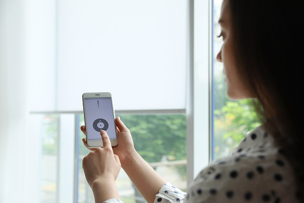 Woman using smart home application on phone to control window blinds indoors